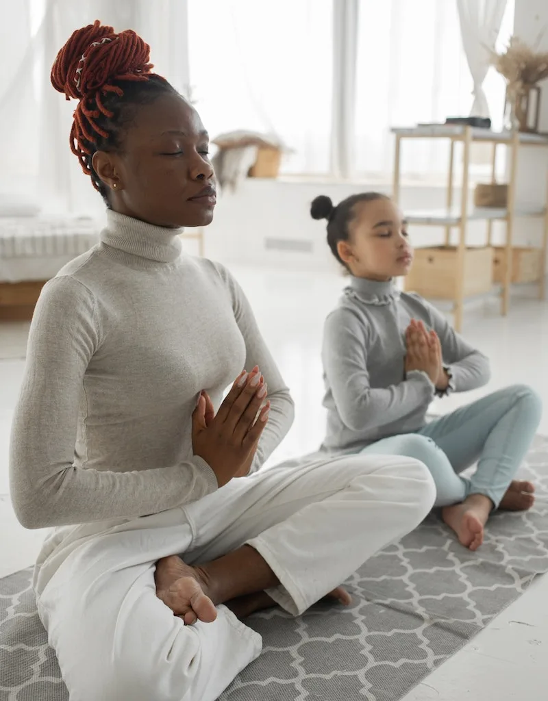 Full body of tranquil young barefooted African American woman in casual clothes sitting on carpet in Padmasana pose with closed eyes while meditating with adorable little daughter at home